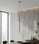 Lampa wisząca Selter 1xG9 w kolorze mokka, wąski, cylindryczny kształt, zawieszona na cienkim przewodzie. Umieszczona w nowoczesnym wnętrzu z betonową ścianą i drewnianym stołem, obok dekoracyjne wazony.