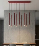 Lampa wisząca Selter 9xG9 w kolorze burgundowym, składająca się z dziewięciu smukłych, pionowych rur zawieszonych na cienkich linkach. Oświetlenie skierowane ku dołowi, montowane na prostokątnej podstawie. W tle betonowa ściana i drewniany stół.