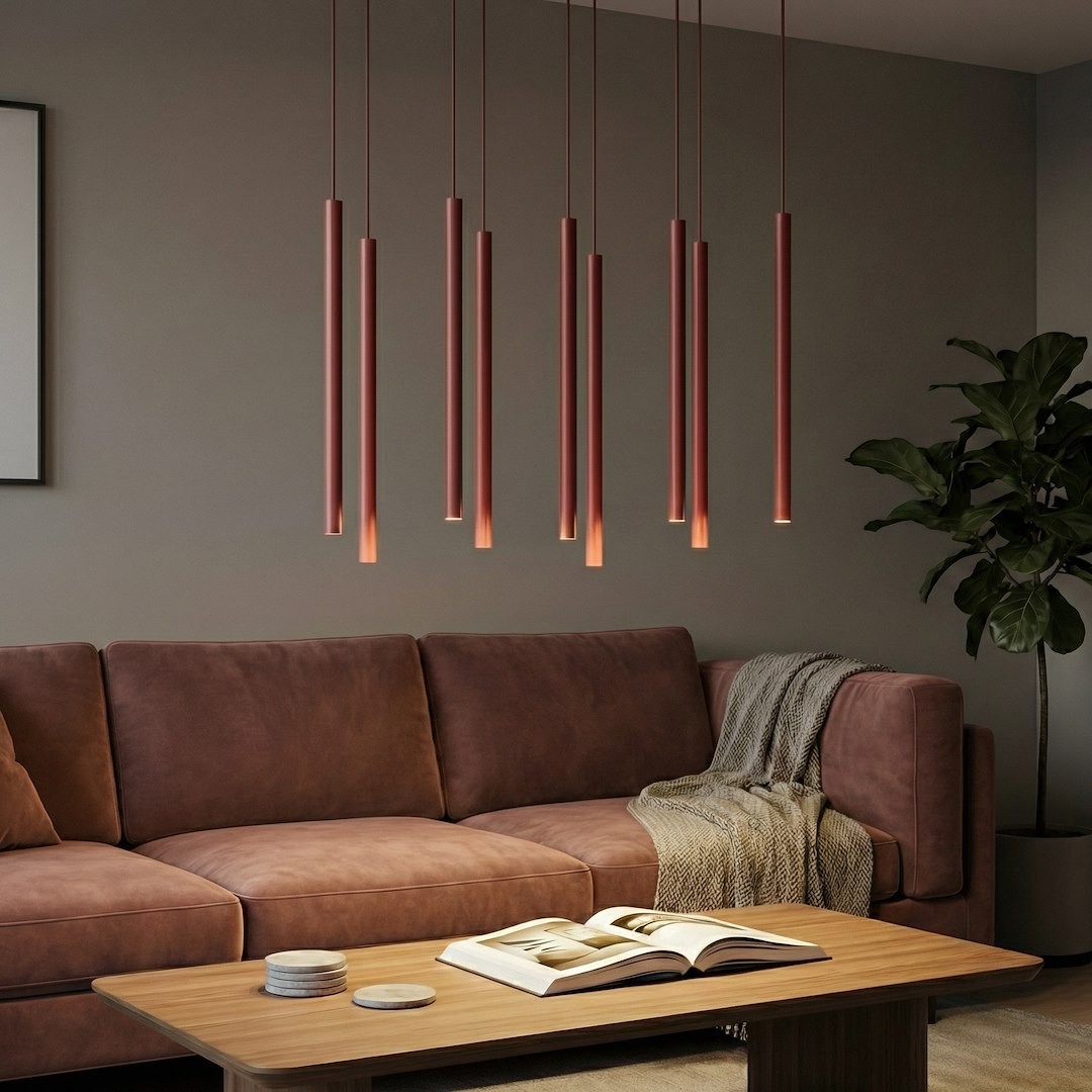 Lampa wisząca Selter 9xG9 w kolorze burgundowym z długimi, cylindrycznymi kloszami zawieszona nad brązową kanapą. W tle widoczne szare ściany, drewniany stół z książką oraz roślina w doniczce.