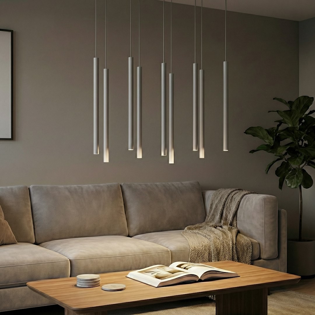 Lampa wisząca Selter 9xG9 w kolorze szarym z długimi, cylindrycznymi kloszami, zawieszona nad sofą w nowoczesnym wnętrzu. W tle widoczna roślina doniczkowa oraz drewniany stół z książką i dekoracjami.