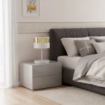 Lampa stołowa Lux E27 w kolorze białym z złotym abażurem, umieszczona na szarej szafce nocnej obok łóżka. Abażur ma perforowaną strukturę, a lampa stoi na okrągłej podstawie. Tło w jasnych odcieniach, z elementami dekoracyjnymi na ścianie.