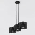 Lampa wisząca Lux w kolorze czarnym z trzema kloszami w kształcie cylindrów, wykonanymi z perforowanego metalu. Zawieszona na dwóch cienkich linkach, z widocznymi żarówkami E27. Proste tło w jasnym kolorze.