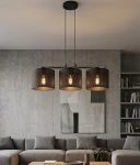 Lampa wisząca Lux z trzema kloszami E27 w kolorze czarnym, zawieszona na długich linkach. Klosze z perforowanej metalowej siatki, emitujące ciepłe światło. Lampa umieszczona nad sofą w nowoczesnym wnętrzu z szarymi ścianami i minimalistycznym wystrojem.