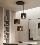 Lampa wisząca Lux 3xE27 w kolorze czarnym, z trzema cylindrycznymi kloszami z perforowanej stali, zawieszona na długich linkach. Widoczna w nowoczesnym wnętrzu z drewnianymi elementami i minimalistycznymi dekoracjami.