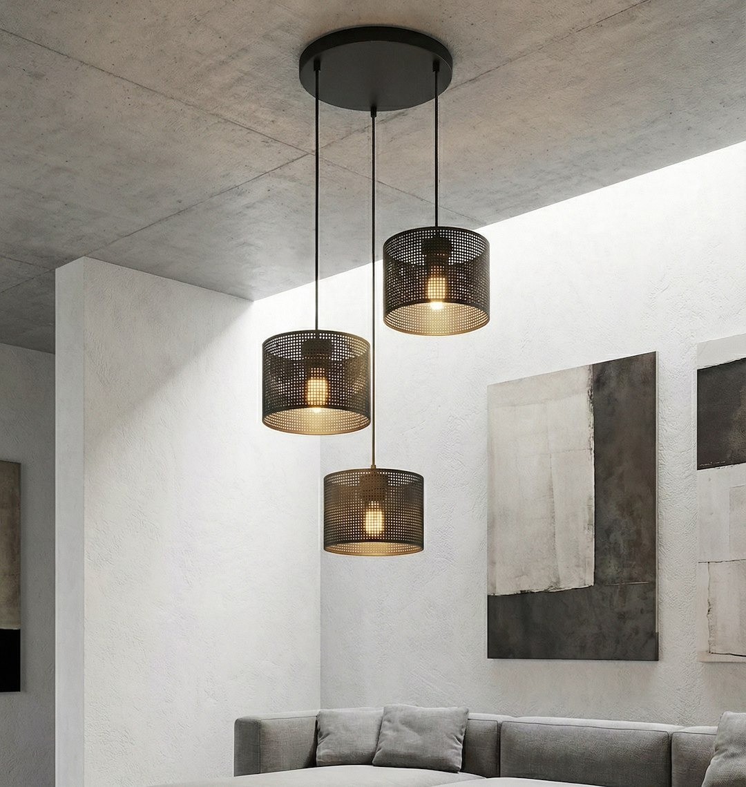 Lampa wisząca Lux 3xE27 w kolorze czarnym z trzema okrągłymi kloszami w metalowej siatce, zawieszona na długich linkach. Zainstalowana na sufitowej podstawie, w nowoczesnym wnętrzu z szarym narożnikiem i abstrakcyjnymi obrazami na ścianach.
