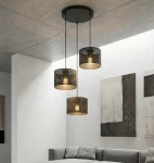 Lampa wisząca Lux 3xE27 w kolorze czarnym z trzema okrągłymi kloszami w metalowej siatce, zawieszona na długich linkach. Zainstalowana na sufitowej podstawie, w nowoczesnym wnętrzu z szarym narożnikiem i abstrakcyjnymi obrazami na ścianach.