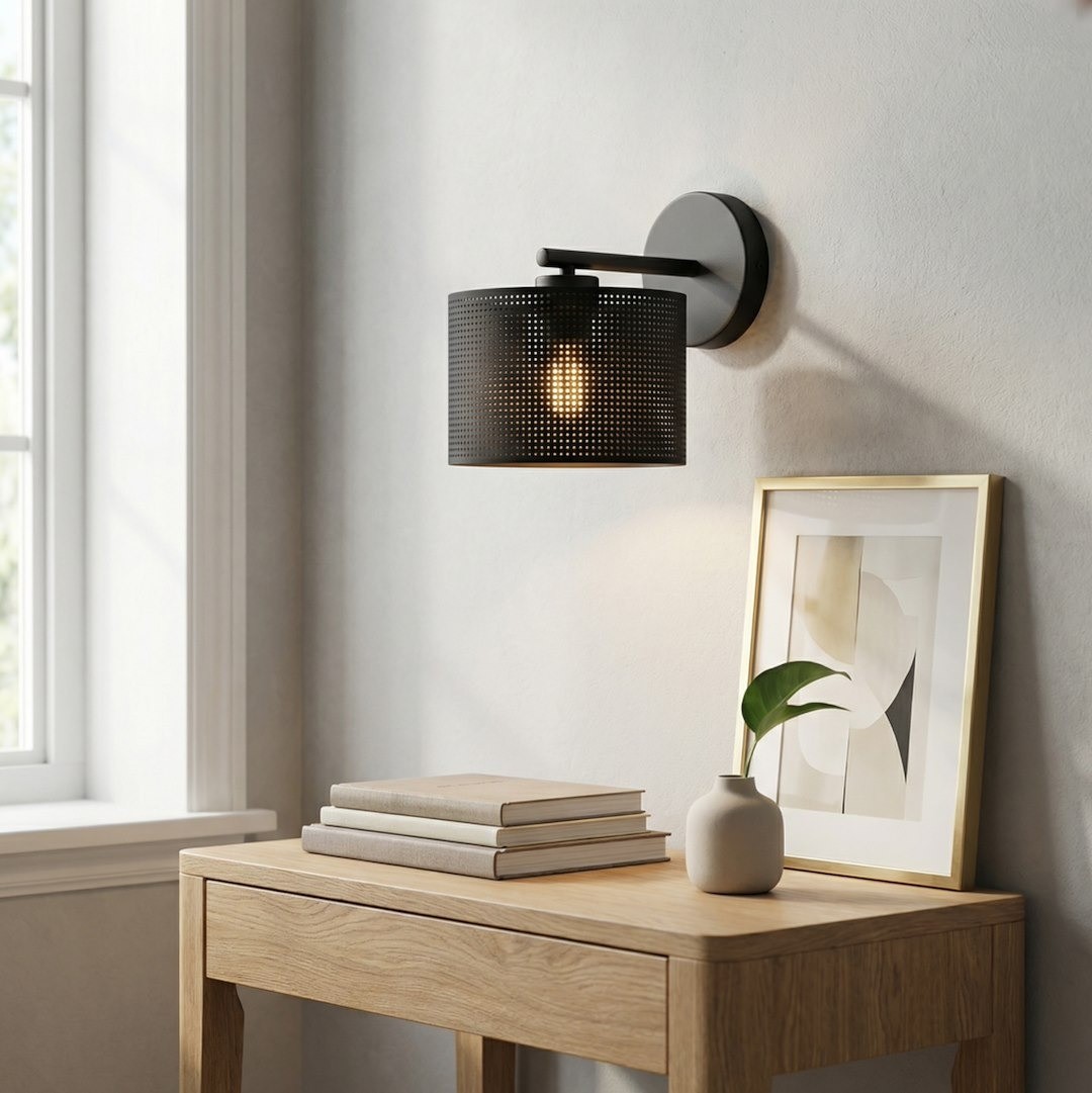 Kinkiet Lux E27 w kolorze czarnym, z metalowym kloszem w perforacje, zamontowany na ścianie. W tle drewniany stół z książkami, ceramiczna waza z rośliną oraz ramka z grafiką. Oświetlenie delikatnie podkreśla detale otoczenia.