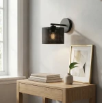 Kinkiet Lux E27 w kolorze czarnym, z metalowym kloszem w perforacje, zamontowany na ścianie. W tle drewniany stół z książkami, ceramiczna waza z rośliną oraz ramka z grafiką. Oświetlenie delikatnie podkreśla detale otoczenia.