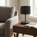 Lampka stołowa Lux E27 w kolorze czarnym, z metalowym kloszem o perforowanej strukturze. Ustawiona na drewnianym stoliku obok szarego fotela, w tle okno. Widoczne książka i okulary na stole.