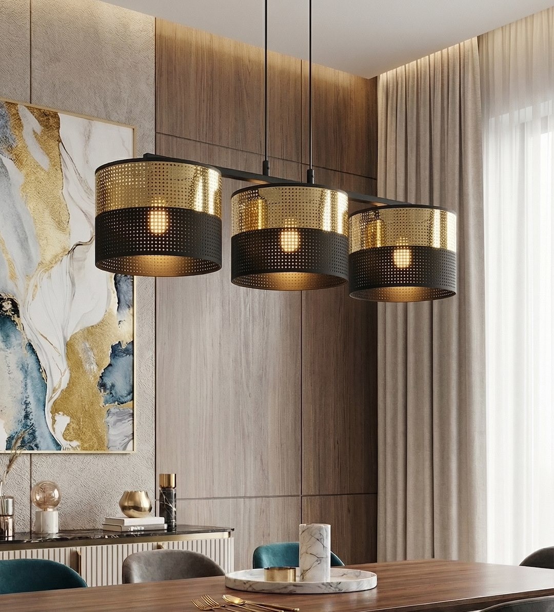 Lampa wisząca Lux w kolorze czarnym z trzema złotymi kloszami, wyposażona w trzy gniazda E27. Ujęcie z dołu, pokazujące lampę zawieszoną nad drewnianym stołem, z nowoczesnym tłem w postaci obrazu i zasłon.
