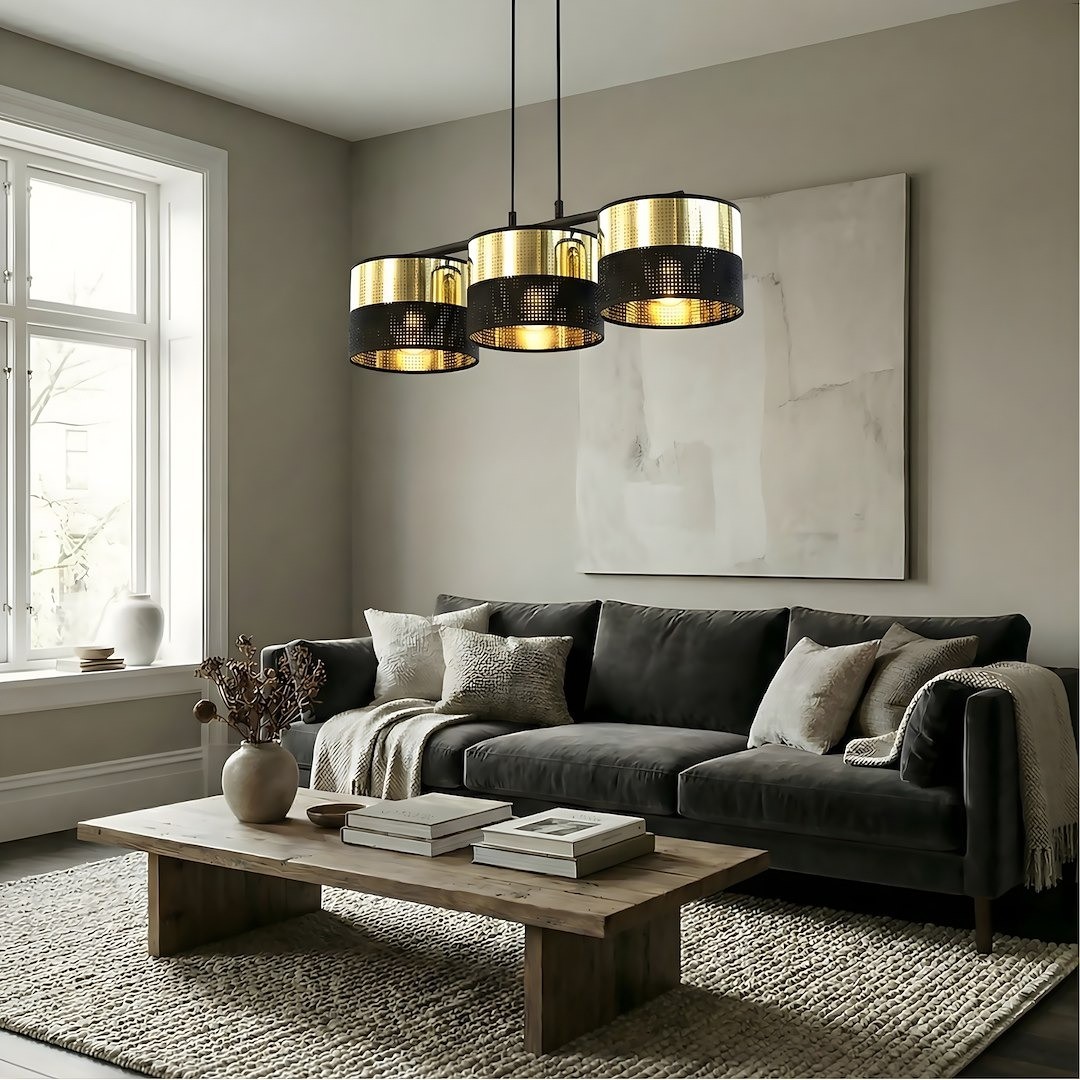 Lampa wisząca Lux 3xE27 w kolorze czarnym i złotym, z trzema cylindrycznymi kloszami, zawieszona na długich linkach. Widoczna w nowoczesnym wnętrzu, nad drewnianym stolikiem w salonie z szaro-beżowym tłem.