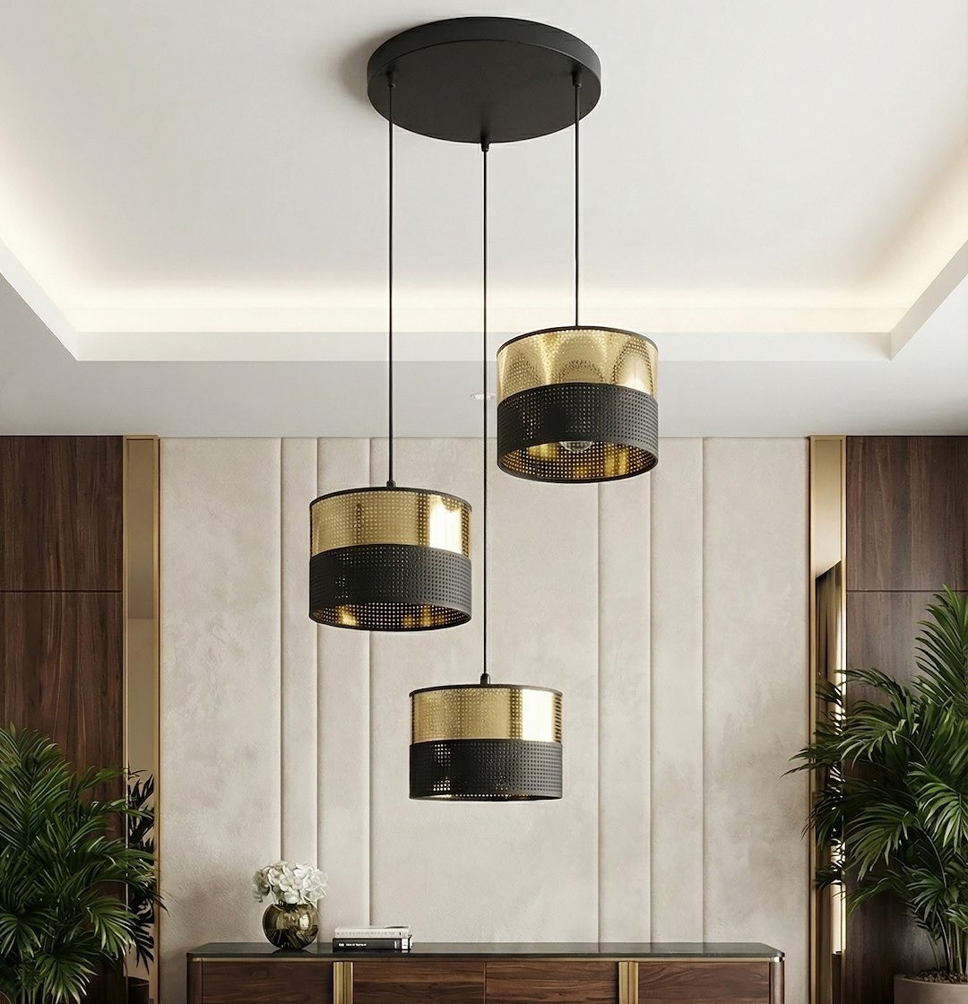 Lampa wisząca Lux 3xE27 w kolorze czarnym i złotym, z trzema cylindrycznymi kloszami. Klosze mają perforowaną teksturę, zawieszone na cienkich przewodach. Produkt montowany sufitowo, w nowoczesnym wnętrzu z drewnianymi akcentami.