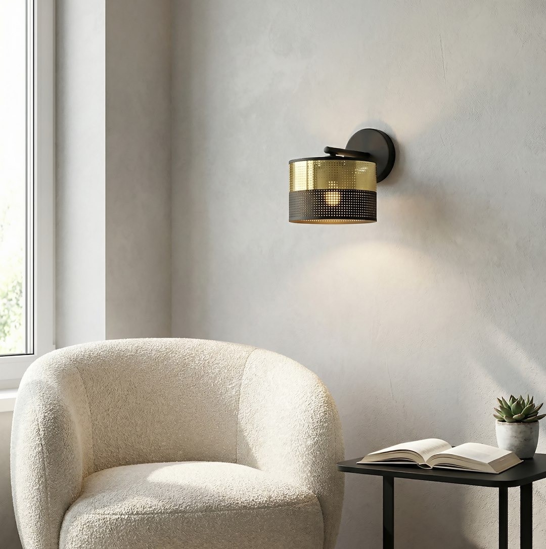 Kinkiet Lux E27 w czarnym i złotym kolorze, z okrągłym kloszem z perforowanej metalu. Montowany na ścianie, z delikatnym światłem. W tle widoczna jasna, miękka sofa i stół z książką oraz doniczką.