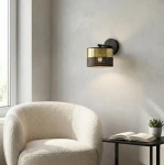 Kinkiet Lux E27 w czarnym i złotym kolorze, z okrągłym kloszem z perforowanej metalu. Montowany na ścianie, z delikatnym światłem. W tle widoczna jasna, miękka sofa i stół z książką oraz doniczką.