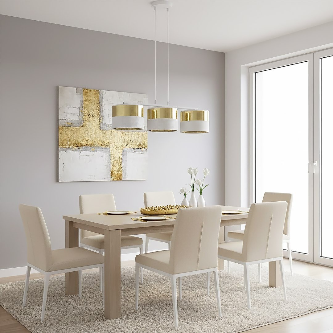 Lampa wisząca Lux 3xE27 w białym kolorze z złotymi akcentami, zawieszona nad drewnianym stołem w jasnej jadalni. Widoczne trzy okrągłe klosze, minimalistyczny design. Tło z szarymi ścianami i dużymi oknami.