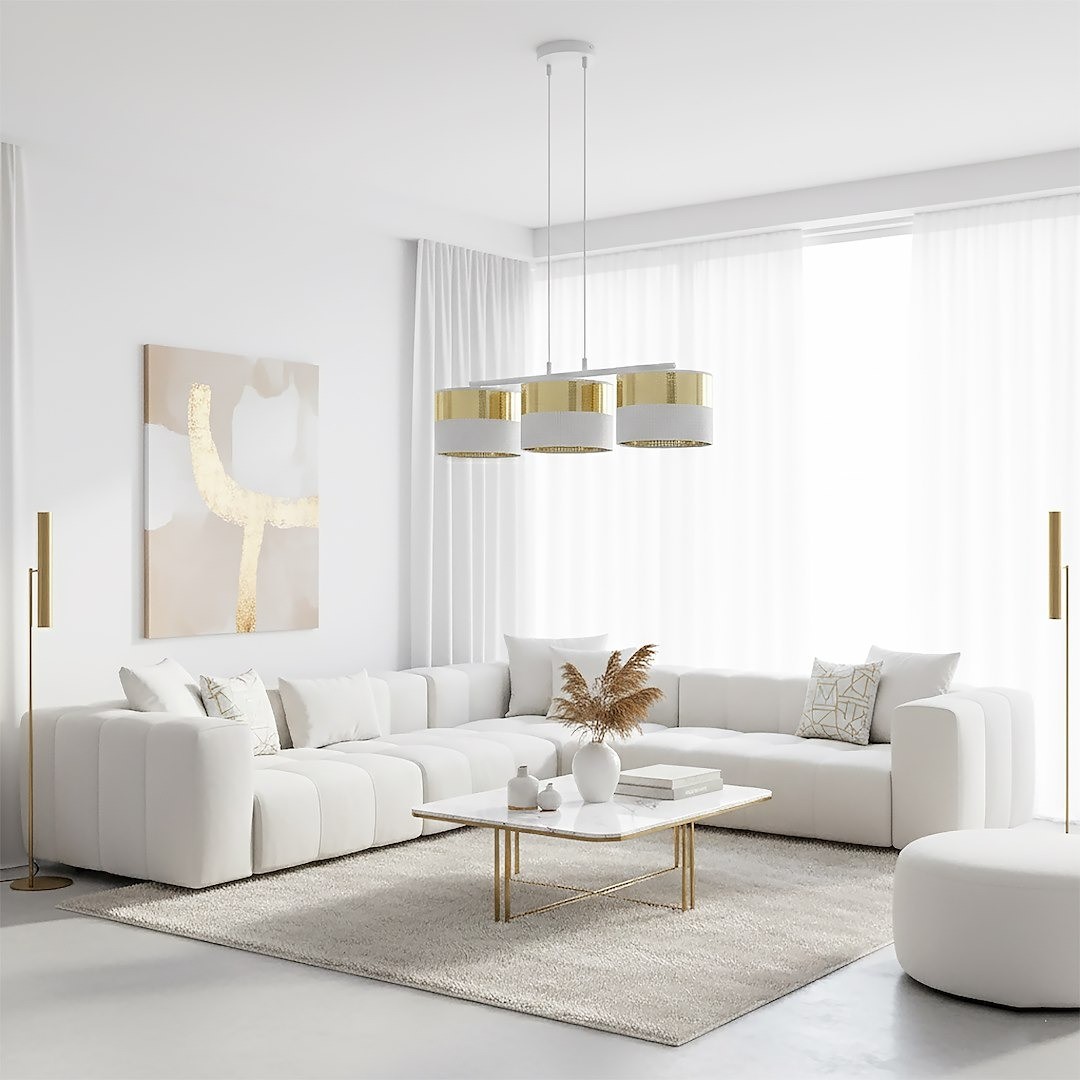 Lampa wisząca Lux 3xE27 w kolorze białym z złotymi detalami, zawieszona w nowoczesnym wnętrzu. Składa się z trzech cylindrycznych kloszy, umieszczonych na cienkich linkach. W tle widoczna jasna kanapa i stół kawowy.