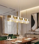 Lampa wisząca Lux 4xE27 w białym kolorze z elementami złotymi, składająca się z czterech okrągłych kloszy. Ujęcie z góry, zawieszona nad drewnianym stołem w eleganckiej aranżacji. Tło w odcieniach beżu i bieli z dekoracjami na ścianach.