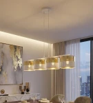 Lampa wisząca Lux 4xE27 w białym kolorze z złotymi elementami, składająca się z czterech okrągłych kloszy. Ujęcie z przodu, z zawieszeniem na białych przewodach. Tło jasne, z dekoracyjną ścianą i zasłonami w tle, tworząc elegancką aranżację.