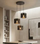 Czarna lampa wisząca Karbon z trzema żarówkami E27, z metalowymi kloszami w siatkowym wzorze. Zawieszona na długich przewodach, umiejscowiona nad stołem w nowoczesnym wnętrzu. Widoczne drewniane elementy oraz minimalistyczne dekoracje w tle.