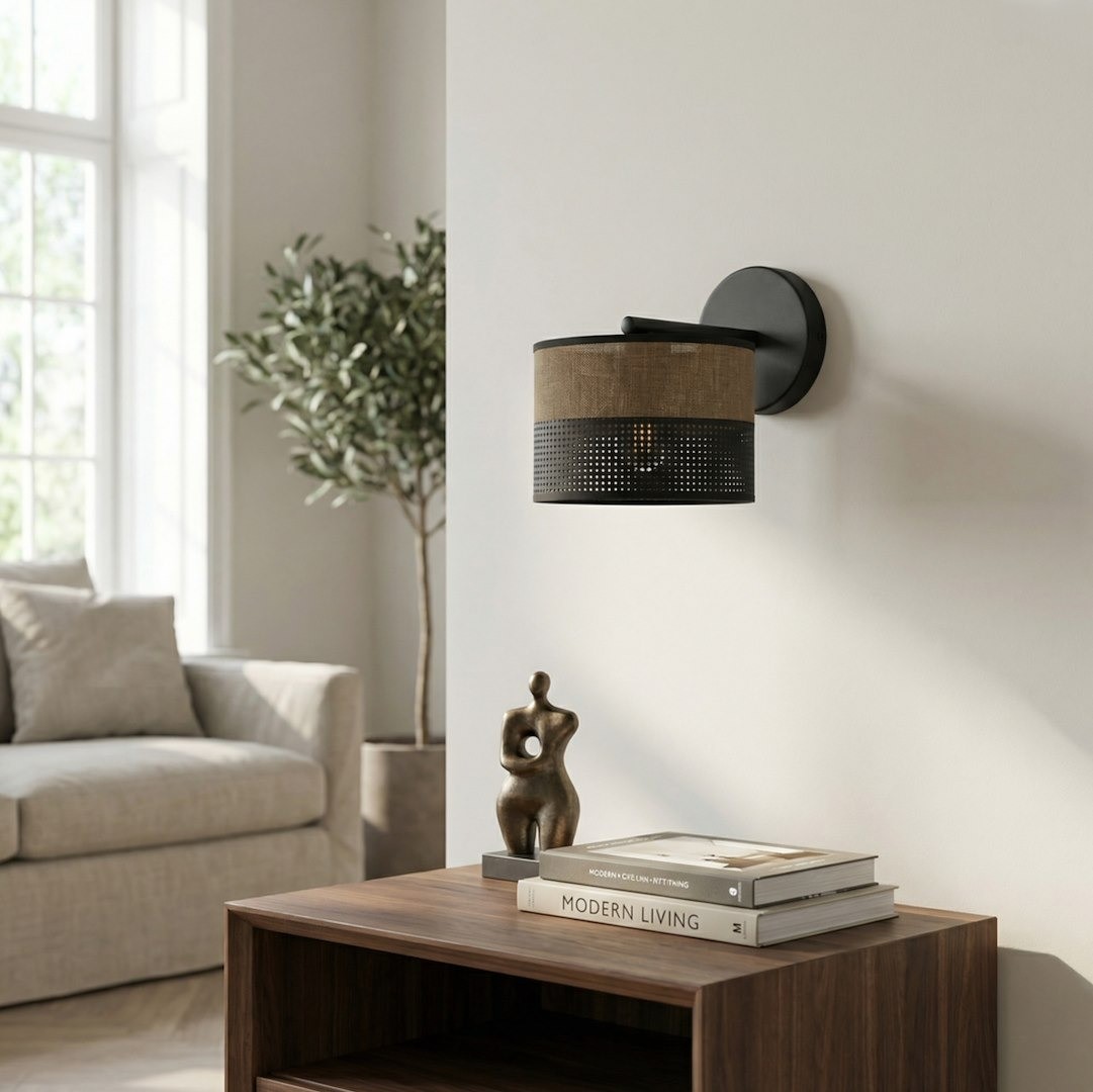 Kinkiet Karbon E27 w kolorze czarnym i brązowym, z okrągłym abażurem z tkaniny i perforowanym dolnym paskiem. Zamontowany na ścianie, w tle widoczny stolik z książkami i figura dekoracyjna, w jasnym wnętrzu.