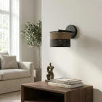 Kinkiet Karbon E27 w kolorze czarnym i brązowym, z okrągłym abażurem z tkaniny i perforowanym dolnym paskiem. Zamontowany na ścianie, w tle widoczny stolik z książkami i figura dekoracyjna, w jasnym wnętrzu.