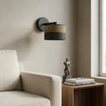Kinkiet Karbon E27 w kolorze czarnym i brązowym, z okrągłym kloszem z perforowanej tkaniny, zamontowany na ścianie obok jasnej sofy. W tle widoczna figura dekoracyjna i stos książek na drewnianym stoliku.