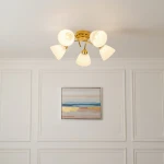 Złota lampa sufitowa Clara z pięcioma kloszami E27 w kształcie stożków, z białym szkłem. Ujęcie z dołu, lampa zamontowana na białym suficie, w tle widoczny obraz w ramie.