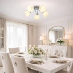 Lampa sufitowa Clara z pięcioma kloszami E27 w kolorze chromowanym. Klosze w kształcie kulistym, biały kolor szkła, umieszczone na metalowej podstawie. Produkt zamontowany na suficie, w eleganckim wnętrzu z białym stołem i kwiatami.
