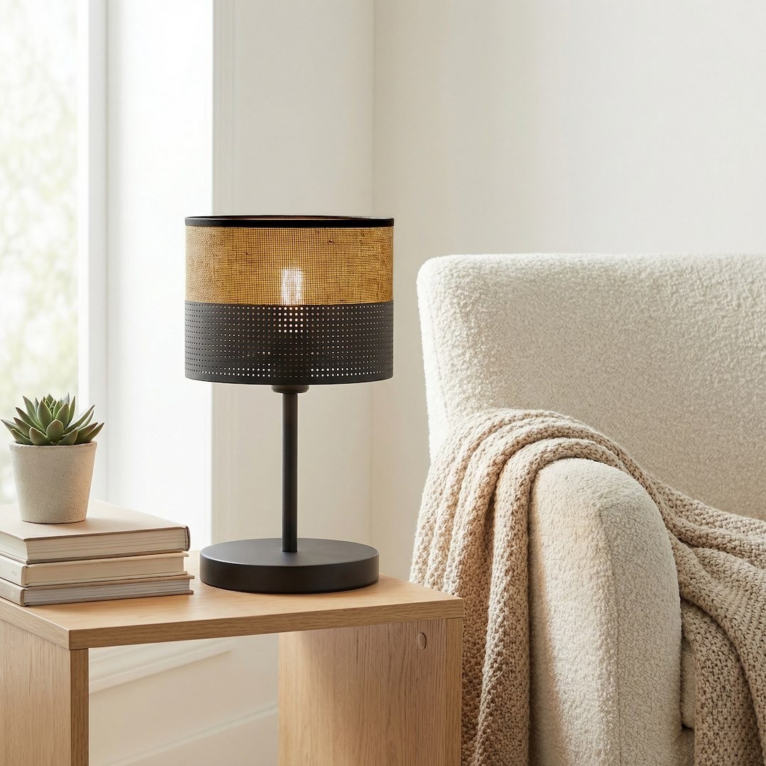 Lampka stołowa Karbon E27 w kolorze czarnym i brązowym, z cylindrycznym kloszem z perforowanego metalu i naturalnego materiału. Ustawiona na drewnianym stoliku obok stosu książek i sukulentu, z widocznym fotelem w tle.
