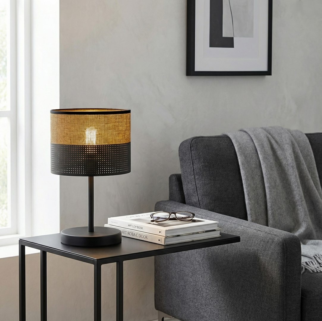 Czarna lampka stołowa E27 z brązowym kloszem, o cylindrycznym kształcie, umieszczona na metalowej podstawie. Dodatki to książki i okulary na stoliku obok szarej sofy. Tło jasne, minimalistyczne.