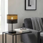 Czarna lampka stołowa E27 z brązowym kloszem, o cylindrycznym kształcie, umieszczona na metalowej podstawie. Dodatki to książki i okulary na stoliku obok szarej sofy. Tło jasne, minimalistyczne.
