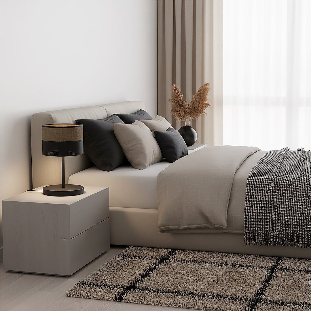 Czarna lampka stołowa E27 z brązowym abażurem, stojąca na białej szafce nocnej obok łóżka. W tle jasno wykończona sypialnia z poduszkami w odcieniach szarości oraz rośliną w doniczce. Wnętrze jasne, z dużymi oknami i zasłonami.