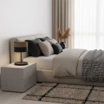 Czarna lampka stołowa E27 z brązowym abażurem, stojąca na białej szafce nocnej obok łóżka. W tle jasno wykończona sypialnia z poduszkami w odcieniach szarości oraz rośliną w doniczce. Wnętrze jasne, z dużymi oknami i zasłonami.