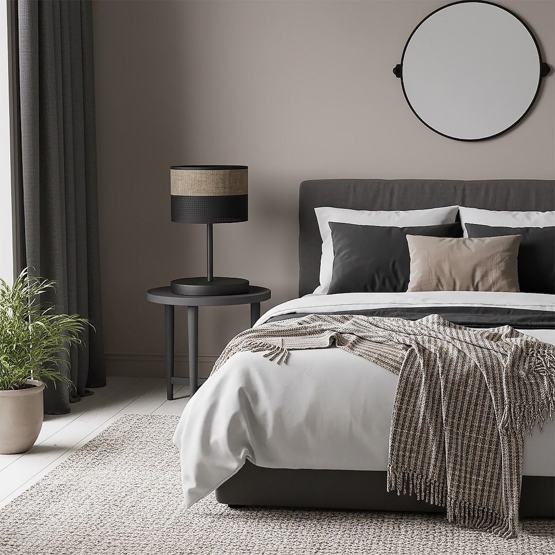 Lampka stołowa Karbon E27 w kolorze czarnym z brązowym abażurem, umieszczona na okrągłym stoliku obok łóżka w nowoczesnej sypialni. Tło w neutralnych odcieniach, z rośliną w donicy oraz dekoracyjnym lustrem na ścianie.