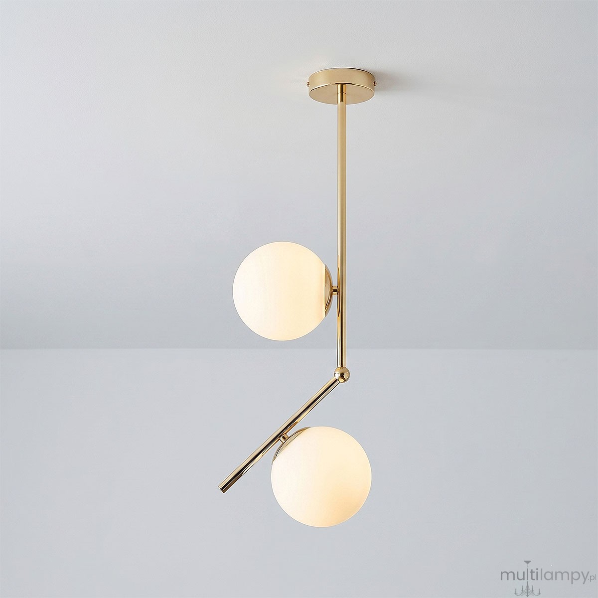 Lampa wisząca Liberty 2-punktowa w kolorze złotym z dwiema białymi, okrągłymi kloszami. Konstrukcja z metalowym ramieniem, wisi w pozycji rozłożonej. Tło w odcieniu szaro-srebrnym.