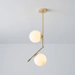 Lampa wisząca Liberty 2-punktowa w kolorze złotym z dwiema białymi, okrągłymi kloszami. Konstrukcja z metalowym ramieniem, wisi w pozycji rozłożonej. Tło w odcieniu szaro-srebrnym.