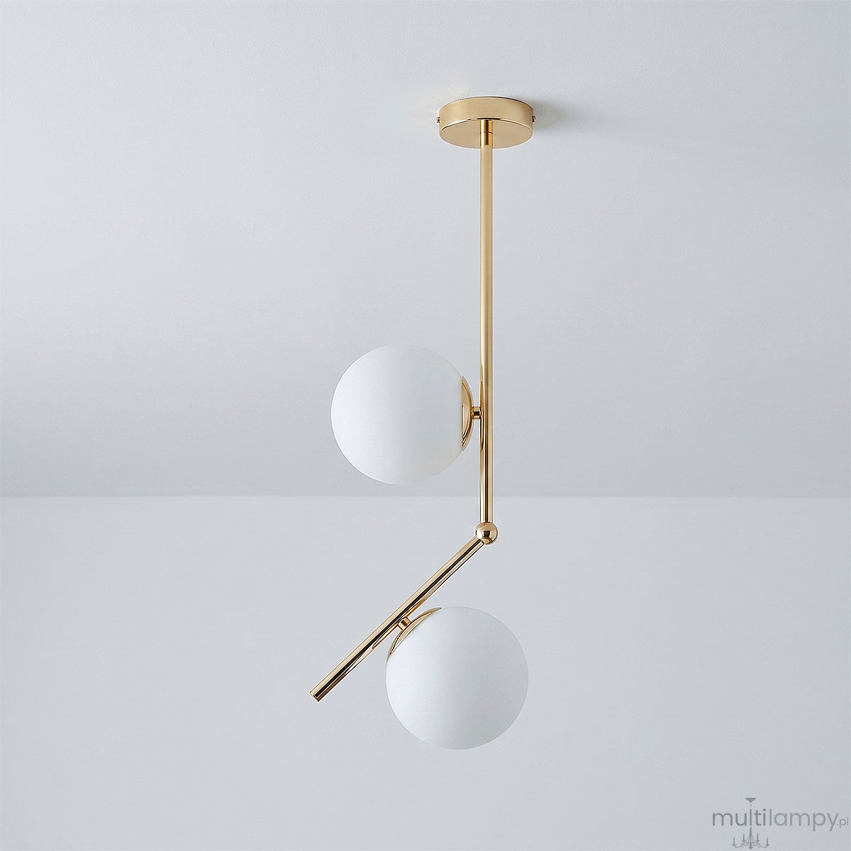 Lampa wisząca Liberty 2-punktowa w kolorze złotym z dwiema białymi, okrągłymi kloszami. Konstrukcja z metalowym ramieniem, wisi w pozycji rozłożonej. Tło w odcieniu szaro-srebrnym.