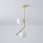 Lampa wisząca Liberty 2-punktowa w kolorze złotym z dwiema białymi, okrągłymi kloszami. Konstrukcja z metalowym ramieniem, wisi w pozycji rozłożonej. Tło w odcieniu szaro-srebrnym.
