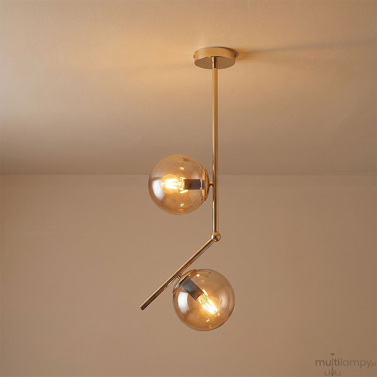 Lampa wisząca Liberty 2-punktowa w kolorze złotym, z dwiema kulistymi kloszami z przezroczystego szkła. Ułożona w pionie, z metalowym, błyszczącym wykończeniem oraz nachyloną rurką, zawieszona na okrągłym stelażu. Tło jasne, neutralne.