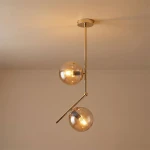 Lampa wisząca Liberty 2-punktowa w kolorze złotym, z dwiema kulistymi kloszami z przezroczystego szkła. Ułożona w pionie, z metalowym, błyszczącym wykończeniem oraz nachyloną rurką, zawieszona na okrągłym stelażu. Tło jasne, neutralne.