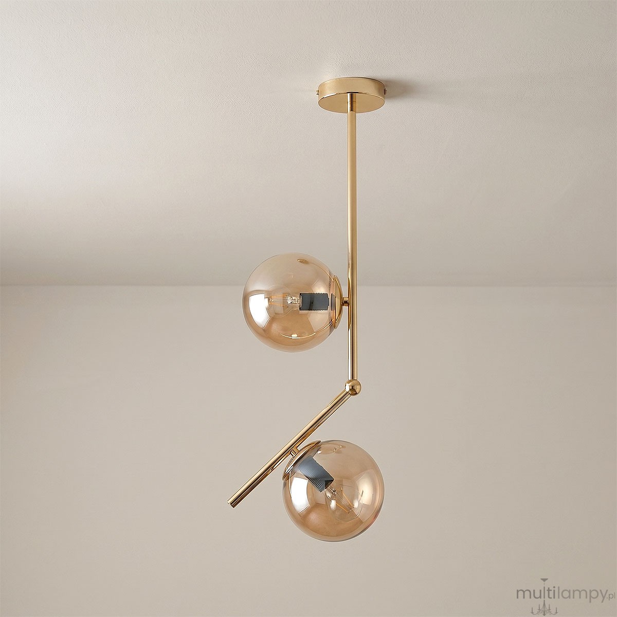 Lampa wisząca Liberty 2-punktowa w kolorze złotym, z dwiema kulistymi kloszami z przezroczystego szkła. Ułożona w pionie, z metalowym, błyszczącym wykończeniem oraz nachyloną rurką, zawieszona na okrągłym stelażu. Tło jasne, neutralne.