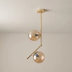 Lampa wisząca Liberty 2-punktowa w kolorze złotym, z dwiema kulistymi kloszami z przezroczystego szkła. Ułożona w pionie, z metalowym, błyszczącym wykończeniem oraz nachyloną rurką, zawieszona na okrągłym stelażu. Tło jasne, neutralne.