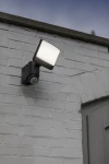 Kinkiet elewacyjny solarny Sunshine w kolorze ciemnoszarym, z kwadratowym kloszem i czujnikiem ruchu, zamontowany na białej ścianie. Produkt jest zamontowany w pozycji stojącej, skierowany na zewnątrz.