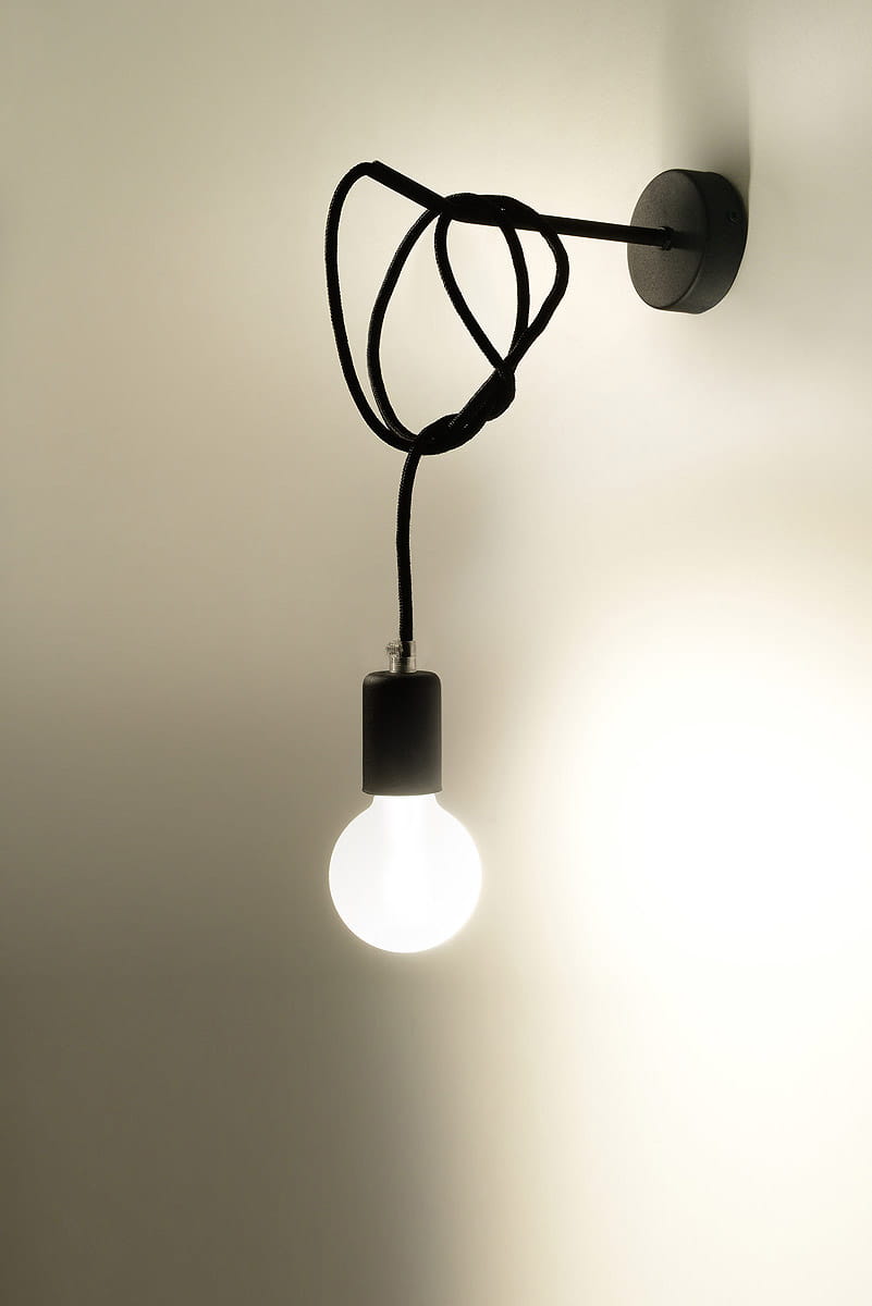 Kinkiet Edison 1-punktowy z czarnym kablem i oprawką. Żarówka w kulistym kształcie emitująca ciepłe światło, zawieszona na ścianie. Prosty, minimalistyczny design, zamontowany na gładkiej, jasnej ścianie.