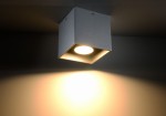 Szara lampa sufitowa 1-punktowa Quad SL.0024 o kwadratowym kształcie, zamontowana na suficie. Wewnątrz znajduje się okrągłe źródło światła, emitujące jasne światło na ścianę w tle. Powierzchnia lampy ma teksturę, co dodaje jej nowoczesnego charakteru.