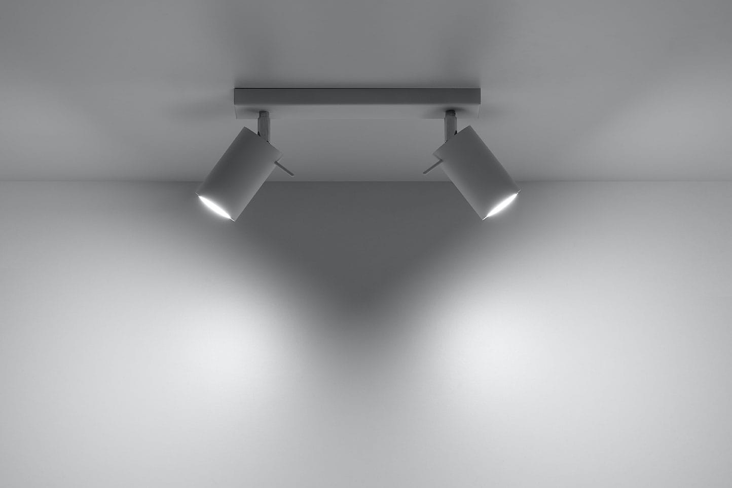 Biała lampa sufitowa typu spot z dwoma punktami świetlnymi, zamontowana na prostokątnej podstawie. Kąt ujęcia z góry, ukazujący dwa cylindryczne klosze skierowane w przeciwnych kierunkach, na jednolitym tle.