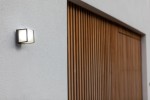 Kinkiet elewacyjny Doblo LED 3000K w ciemnoszarym kolorze, zamontowany na ścianie, prostokątny kształt z białym kloszem, emitujący ciepłe światło. W tle widoczne drewniane deski.