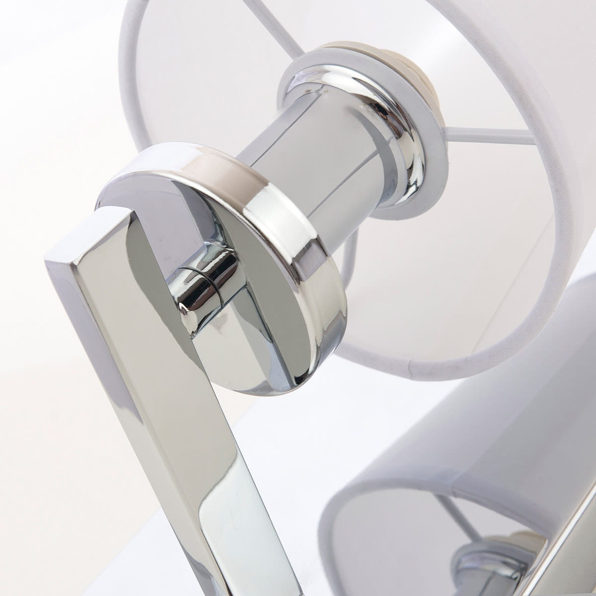 Kinkiet London 1-punktowy w kolorze chromu i białym, z metalową podstawą i cylindrycznym kloszem. Ujęcie z boku, eksponujące połysk metalowych elementów oraz detal mocowania klosza. Tło jasne, neutralne.