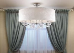 Lampa sufitowa Amsterdam 8-punktowa, okrągły klosz w jasnym kolorze z delikatną fakturą, metalowe elementy w chromowanej barwie, wiszące kryształki. Ujęcie z góry, lampa zawieszona w centralnym punkcie, w tle zasłony w kolorze morskim.