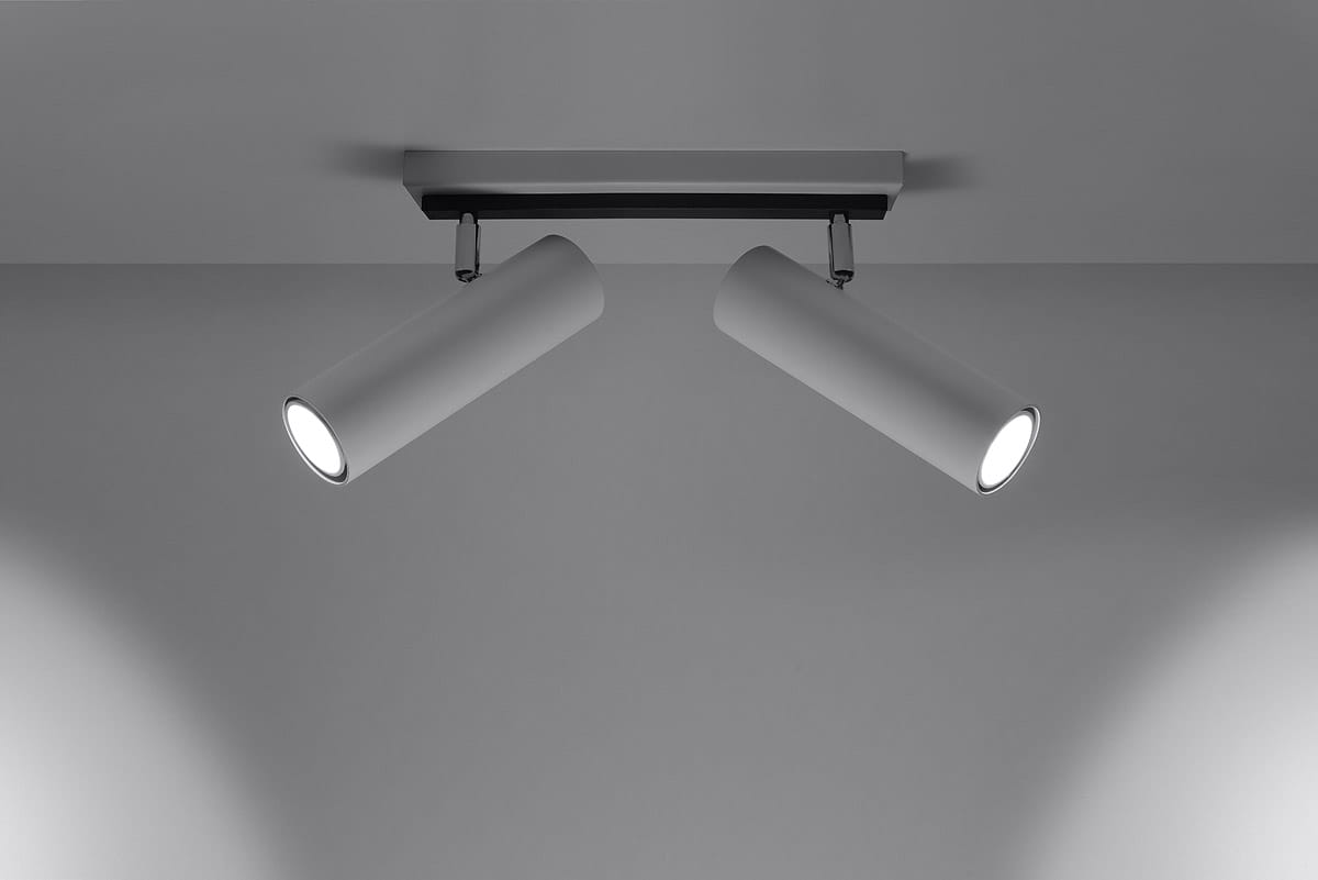 Biała lampa sufitowa Direzione z dwoma cylindrycznymi punktami świetlnymi, zamontowana na prostokątnej podstawie. Ujęcie z dołu, ukazujące symetryczne rozmieszczenie reflektorów, które emitują jasne światło. Tło jednolite, szare.